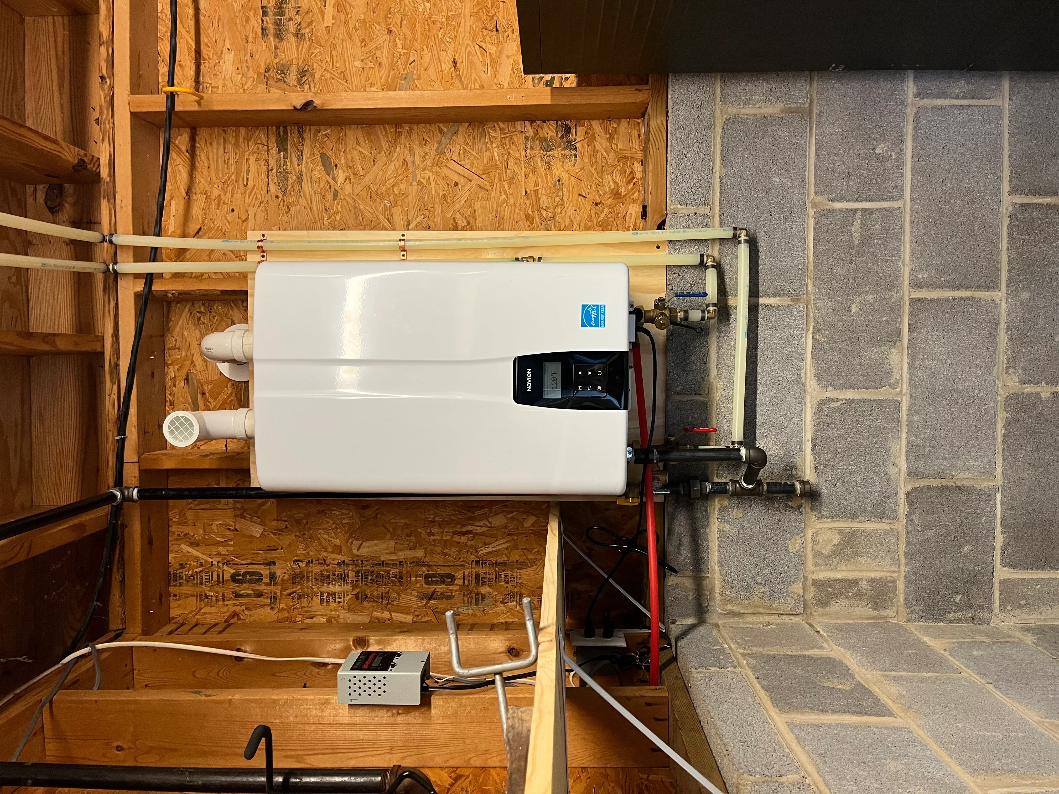 White tankless water heater with digital display mounted on wood-framed wall with copper and PVC piping connections