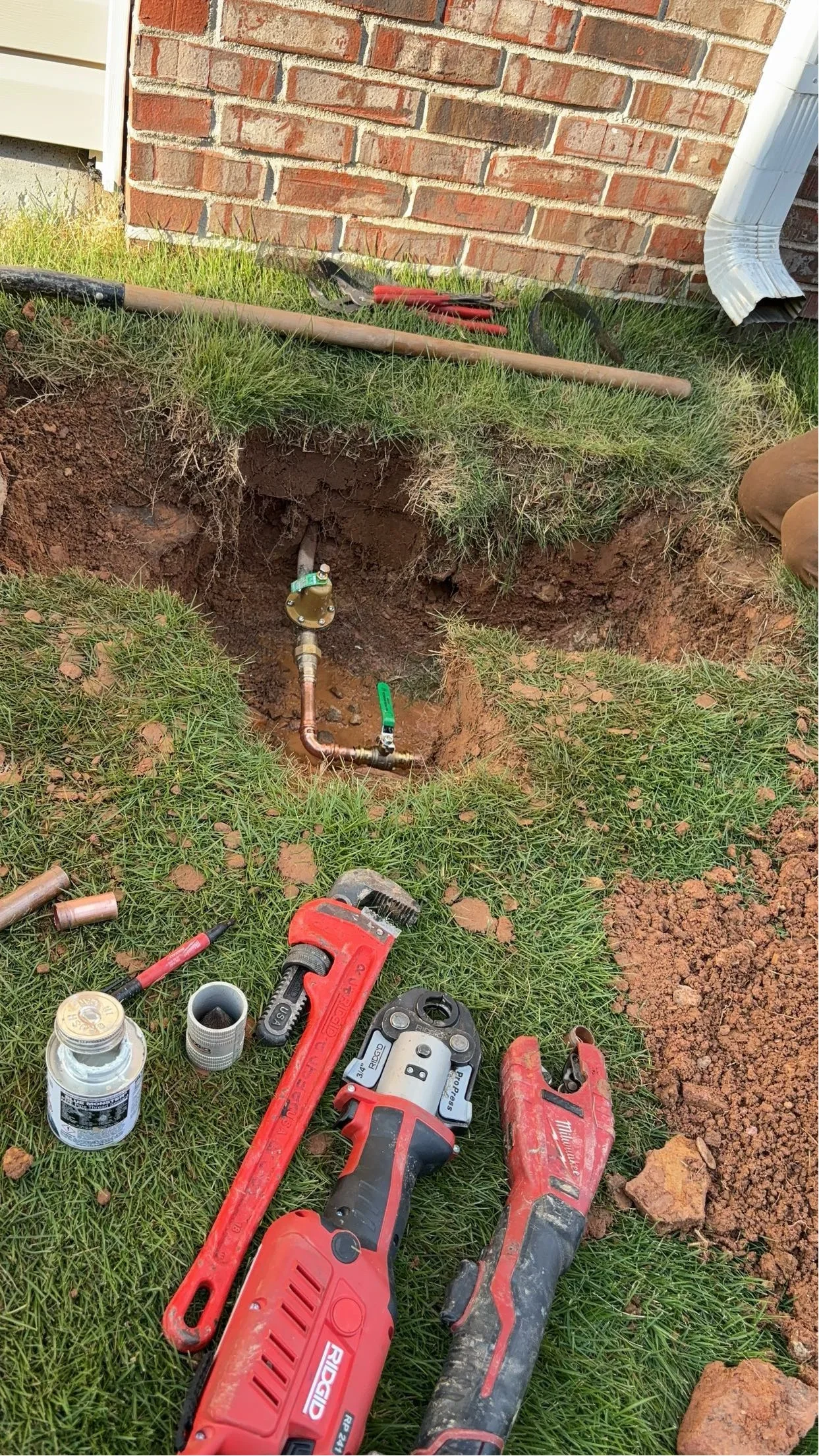 Excavated trench showing copper water line, shutoff valve, and PVC piping installation near brick foundation