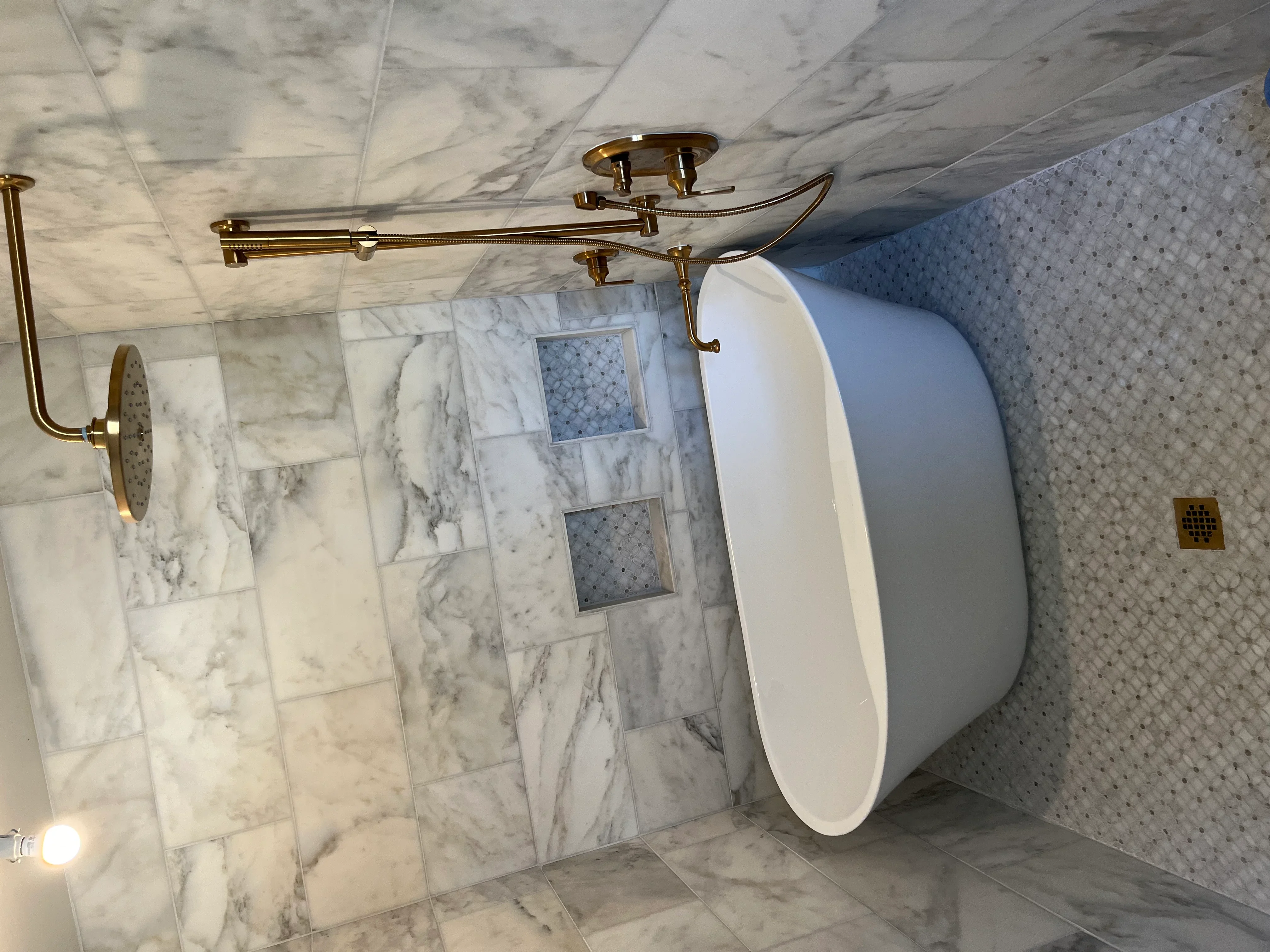 Marble tile bathroom with brass rainfall showerhead, curved supply lines, and freestanding white soaking tub